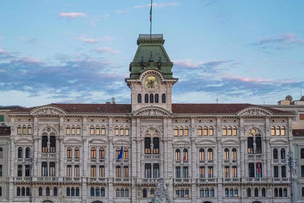 Neoclassical architectural landmark in Trieste, Italy, with clock tower against blue sky at dusk.